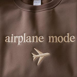 May include: A brown sweatshirt with the words "airplane mode" embroidered in a light tan color. Below the text is a matching embroidered airplane graphic. The sweatshirt has a crew neck and long sleeves.