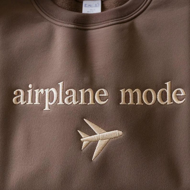 May include: A brown sweatshirt with the words "airplane mode" embroidered in a light tan color. Below the text is a matching embroidered airplane graphic. The sweatshirt has a crew neck and long sleeves.