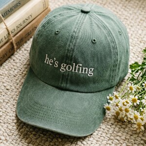 May include: A sage green baseball cap with the embroidered text "he's golfing" in white. The hat has a curved brim and a button on top. The cap is sitting on a textured surface with books in the background.