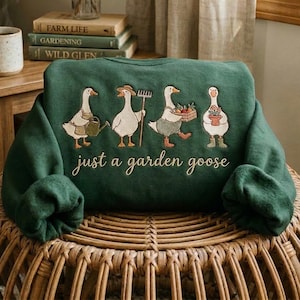 May include: A dark green sweatshirt featuring an embroidered design of four white geese engaged in gardening activities, with the text just a garden goose written in cursive script below the geese. The geese wear hats and carry tools.