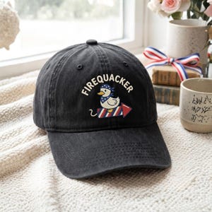 May include: A charcoal gray baseball cap featuring an embroidered graphic of a white duck wearing a blue and white star-patterned bandana, riding a red, white, and blue firework rocket, with the text FIREQUACKER arched above the duck.