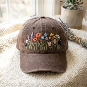 May include: Brown baseball cap with embroidered colorful flowers. The floral design includes orange poppies, blue forget-me-nots, yellow daisies, and lavender sprigs. The cap has a curved brim and a button on top.