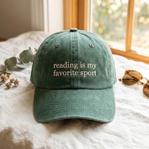 May include: A forest green baseball cap with a distressed fabric finish. The front panel features the embroidered text "reading is my favorite sport" in a light cream color. The cap has a structured crown, a curved brim, and a button on top.