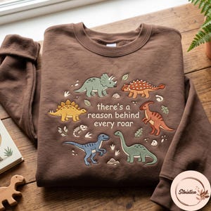 May include: A brown crewneck sweatshirt featuring an embroidered design of five colourful dinosaurs arranged in a circle around the text "there's a reason behind every roar." The dinosaurs include a green triceratops, orange ankylosaurus, yellow stegosaurus, blue raptor, and green long-necked sauropod.