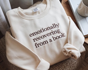 May include: A cream-coloured crewneck sweatshirt featuring dark brown embroidered text on the chest that reads, "emotionally recovering from a book." The garment has long sleeves with ribbed cuffs and a ribbed neckline, showcasing a soft fabric texture.