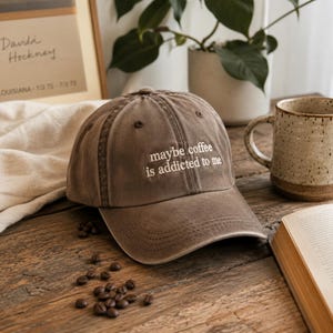 May include: A brown baseball cap with the embroidered text "maybe coffee is addicted to me." The cap is sitting on a wooden surface with coffee beans scattered around it. A ceramic mug and a book are also visible.