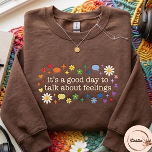 Könnte beinhalten: Ein braunes Sweatshirt mit Rundhalsausschnitt und dem gestickten Text "it's a good day to talk about feelings" in Weiß, umgeben von bunten gestickten Symbolen wie Gänseblümchen, Sprechblasen, Herzen und vierzackigen Sternen.