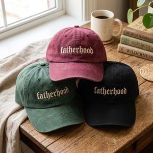 May include: Three baseball caps in burgundy, green, and black, each embroidered with the word "fatherhood" in white script. The caps are displayed on a wooden surface, with a coffee mug and books in the background.