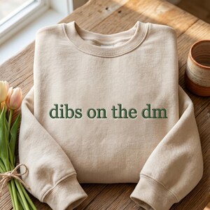 Op de afbeelding: Een beige sweater met ronde hals en de geborduurde tekst "dibs on the dm" in donkergroene draad. Het kledingstuk heeft lange mouwen met geribbelde boorden en een geribbelde halslijn, met een strak en eenvoudig ontwerp in een neutrale kleur.