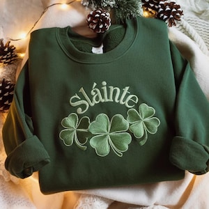 May include: A dark green sweatshirt with the word "Sl&aacute;inte" embroidered in white script above three embroidered shamrocks. The sweatshirt has long sleeves and a crew neck. The image is taken from a high angle.
