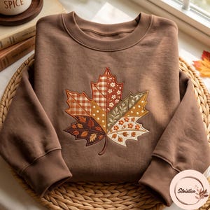 Puede incluir: Una sudadera marrón de cuello redondo con una gran hoja de arce bordada en el pecho. La hoja está dividida en segmentos con varios patrones, incluyendo cuadros vichy, lunares y motivos florales en tonos naranja, crema y marrón.