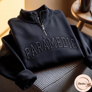 May include: A black quarter-zip sweatshirt featuring a high collar and a silver-toned metal zip. The word PARAMEDIC is embroidered in a matching black thread across the chest in a bold, arched, collegiate-style font with a textured finish.