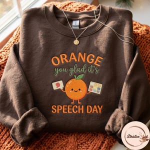 May include: A dark brown crewneck sweatshirt featuring an embroidered orange character holding a speech bubble and a colour palette. The text reads Orange you glad it's SPEECH DAY in orange and green thread. A gold chain necklace rests on the collar.