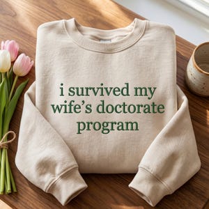 May include: A beige crewneck sweatshirt with the text "i survived my wife's doctorate program" in green lettering. The sweatshirt is displayed on a wooden surface, with a bouquet of tulips and a ceramic mug in the background.