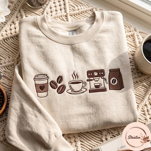 Puede incluir: Una sudadera de cuello redondo color crema con bordados marrones que muestran una taza de café con un corazón, tres granos de café, una taza humeante sobre un plato, una máquina de café expreso y una bolsa de café en el centro.
