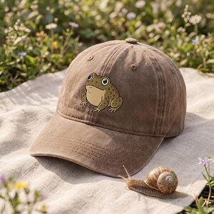 Puede incluir: Una gorra de béisbol marrón con una ilustración gráfica centrada de un sapo verde y beige con ojos grandes. La gorra presenta una corona estructurada, una visera curva y ojales cosidos en los paneles superiores de la tela.