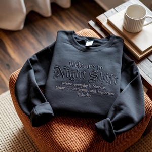 May include: A black crewneck sweatshirt featuring dark embroidery that reads: Welcome to Night Shift, where everyday is Monday, today is yesterday, and tomorrow is today. The garment has long sleeves and a ribbed collar with a white interior tag.