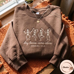 May include: A brown crewneck sweatshirt featuring an embroidered design of four dancing skeletons. Below the skeletons, the text reads "dry bones come alive" in a cursive font, followed by the printed text "EZEKIEL 37:5" in a sans-serif font.