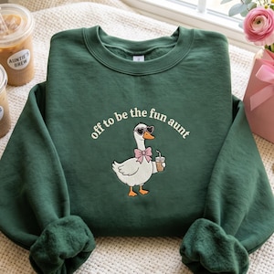 May include: A dark green crewneck sweatshirt featuring a graphic of a white goose wearing heart-shaped sunglasses and a pink bow tie while holding a coffee cup. The text "off to be the fun aunt" is printed in a cream-coloured font above the goose.