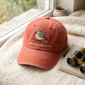 Puede incluir: Gorra de béisbol de color óxido con un diseño de pájaro bordado. La gorra presenta un pájaro azul, amarillo y negro posado en una rama, con el texto "easily distracted by birds" debajo. La gorra tiene una visera curva y un botón en la parte superior.