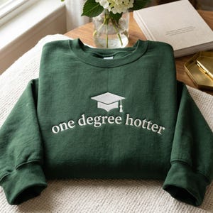May include: A dark green sweatshirt with white embroidered text and a graduation cap design. The text reads "one degree hotter". The sweatshirt is folded on a white blanket.