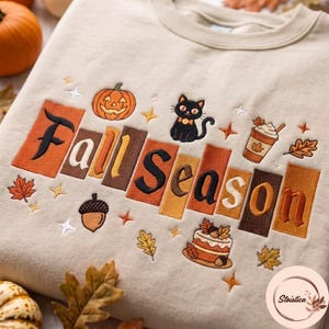 May include: A beige crewneck sweatshirt featuring embroidered text that reads Fall Season. The design includes a jack-o'-lantern, a black cat, a coffee cup, an acorn, a cake, and various autumn leaves and stars in orange, brown, and cream tones.