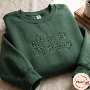 May include: A forest green crewneck sweatshirt featuring the embroidered text "off to do OT things" in a matching dark green thread. The garment has a ribbed collar, cuffs, and hem, with a clean, solid-colored fabric finish throughout the front.
