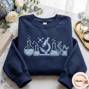 Puede incluir: Una sudadera azul marino de cuello redondo con bordado en azul claro de equipo de laboratorio científico, que incluye un matraz de fondo redondo, un microscopio, un matraz Erlenmeyer, una doble hélice de ADN y una botella pequeña con una hoja.