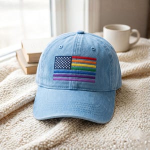 May include: A light blue denim baseball cap featuring an embroidered patch on the front. The patch displays a rectangular design combining the American flag with white stars on a blue field and horizontal rainbow stripes in red, orange, yellow, green, blue, and purple.