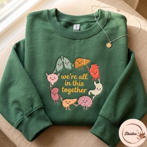 May include: A forest green crewneck sweatshirt featuring a central graphic of cartoon human organs holding hands in a circle. The text "we're all in this together" is printed in yellow in the centre. A thin silver chain necklace rests on top.