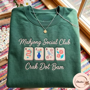 May include: A dark green crewneck sweatshirt featuring embroidered text that reads Mahjong Social Club and Crak Dot Bam. Between the text lines are four mahjong tiles depicting a dragon, a vase with flowers, a bird, and floral patterns.
