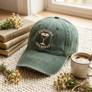 Puede incluir: Gorra de béisbol verde con la inscripción "Espresso Yourself" bordada en blanco. La gorra presenta un gráfico con temática de café y una visera curva. Una taza de café y flores están en el fondo.