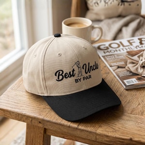 May include: A tan baseball cap with a black brim. The cap features the text "Best Uncle BY PAR" in black script, along with a small graphic of a golfer. A black button sits atop the cap.