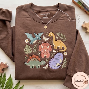 May include: A brown long-sleeve sweatshirt featuring an embroidered graphic of various dinosaurs, including a pterodactyl, triceratops, long-neck, and stegosaurus, surrounded by small flowers, leaves, and butterflies. The inner label reads Cotton Blend S Made in USA.
