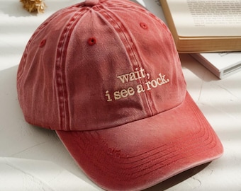 Puede incluir: Gorra de béisbol roja con la frase bordada "wait, i see a rock." La gorra tiene una visera curva y un aspecto vintage descolorido. El sombrero está hecho de una tela suave y desgastada.