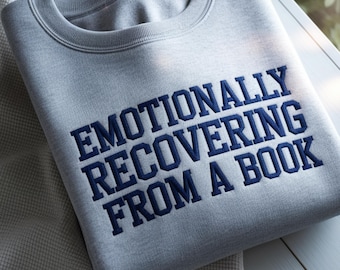 May include: A light grey crewneck sweatshirt featuring dark navy blue embroidered text that reads "EMOTIONALLY RECOVERING FROM A BOOK" in a bold, capitalised, serif font. The garment has a ribbed collar and a smooth, soft-knit fabric texture.