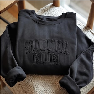 May include: A black crewneck sweatshirt featuring the text SOCCER MOM embroidered in a matching black thread across the chest. The lettering is in a bold, collegiate-style font with an arched layout for the word SOCCER and a straight layout for MOM.