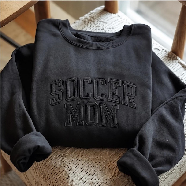 May include: A black crewneck sweatshirt featuring the text SOCCER MOM embroidered in a matching black thread across the chest. The lettering is in a bold, collegiate-style font with an arched layout for the word SOCCER and a straight layout for MOM.