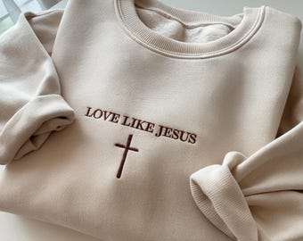 以下が含まれることがあります： 胸元にダークブラウンの刺繍が施されたベージュのクルーネックトレーナー。LOVE LIKE JESUSというテキストがシンプルな十字架のシンボルの上に配置されています。長袖で、袖口と襟元はリブ編みになっており、柔らかな生地で作られています。