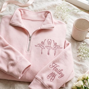 May include: A light pink quarter-zip pullover featuring an embroidered design of three ballerinas on the chest. The left sleeve is embroidered with the name Hailey in cursive script above a pair of ballet slippers in a matching pink thread.