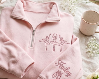 May include: A light pink quarter-zip sweatshirt featuring an embroidered design of three ballet dancers on the chest. The sleeve is embroidered with the name Hailey in cursive script above a pair of ballet slippers in a matching pink thread.