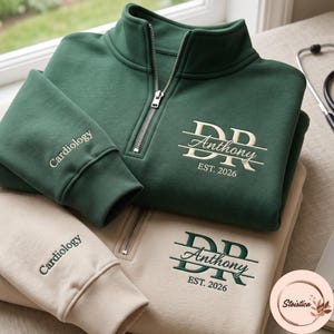May include: A dark green quarter-zip sweatshirt with cream embroidery reading DR Anthony EST. 2026 and Cardiology on the sleeve, stacked on a beige quarter-zip sweatshirt with dark green embroidery reading DR Anthony EST. 2026 and Cardiology on the sleeve.