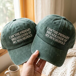 May include: Two forest green baseball caps with a distressed, washed finish. Each cap features white embroidered text that reads: I AM THE PRIMARY BREADLOSER OF MY HOUSEHOLD. The hats have a structured crown, a curved brim, and a button on top.
