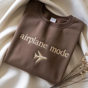 May include: A brown sweatshirt with the words "airplane mode" embroidered in a light beige color, along with a small airplane graphic. The sweatshirt is folded and resting on a cream-colored fabric.