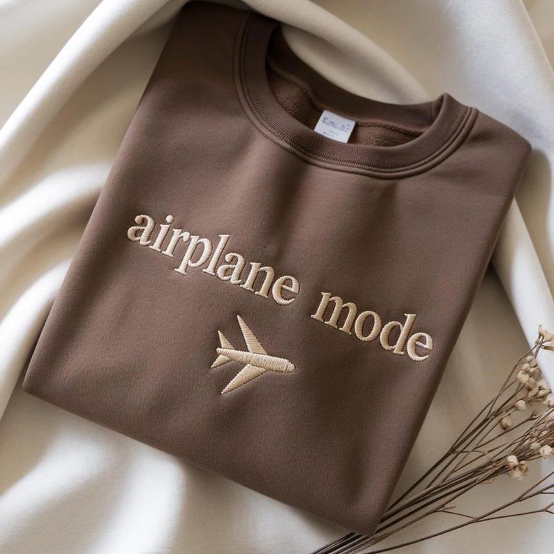 May include: A brown sweatshirt with the words "airplane mode" embroidered in a light beige color, along with a small airplane graphic. The sweatshirt is folded and resting on a cream-colored fabric.