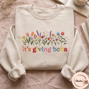 May include: A beige crewneck sweatshirt featuring an embroidered design of colourful wildflowers in shades of pink, orange, purple, yellow, blue, and red. Below the flowers, the text it's giving bcba is embroidered in multi-coloured letters.