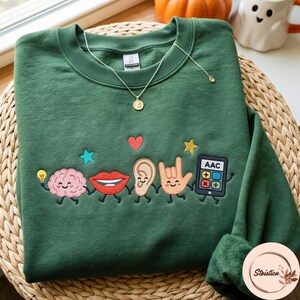May include: A dark green crewneck sweatshirt featuring an embroidered design of five anthropomorphic characters: a pink brain holding a lightbulb, red lips, a tan ear, an orange hand making a sign language gesture, and a black AAC calculator device.