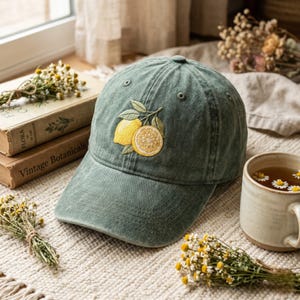 May include: A sage green baseball cap with an embroidered lemon design. The design features two yellow lemons and green leaves. The cap is sitting on a white surface with books and a cup of tea in the background.