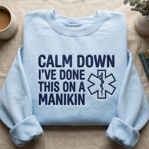 May include: A light blue crewneck sweatshirt featuring dark blue embroidered text that reads CALM DOWN I'VE DONE THIS ON A MANIKIN, accompanied by a dark blue Star of Life medical symbol with a serpent and staff design on the right side.