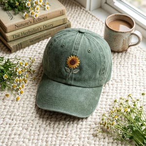 Könnte beinhalten: Grüne Baseballkappe mit gesticktem Sonnenblumenmuster. Die Kappe besteht aus einem weichen, abgenutzten Stoff und liegt auf einer cremefarbenen, strukturierten Oberfläche. Im Hintergrund sind alte Bücher und eine Tasse Kaffee zu sehen.
